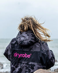 Woman stood on beach with back to camera, wearing dryrobe Advance Long Sleeve REMIX Range change poncho in Black Camo Pink Black