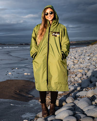 Woman stood on the beach in the sun, wearing dryrobe Advance Long Sleeve REMIX Range change poncho in Forest Green Black Camo