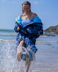 Woman on a beach splashing through shoreline, wearing swimsuit underneath dryrobe Advance Long Sleeve changing poncho in Blue Camo Blue