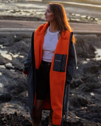 Woman stood in sun on beach, wearing dryrobe Advance Long Sleeve change poncho in Dark Grey Orange