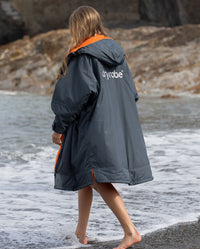 Girl walking along shore line on the beach wearing dryrobe AdvanceLong Sleeve Change poncho in Dark Grey Orange