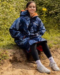 Girl sat on grass by hedge, wearing dryrobe Advance Long Sleeve Changing poncho in Blue Camo Orange