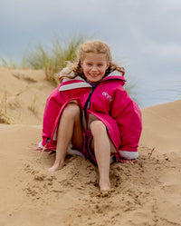 Girl sitting on sand, wearing dryrobe Advance Long Sleeve Changing poncho in Pink Light Grey 