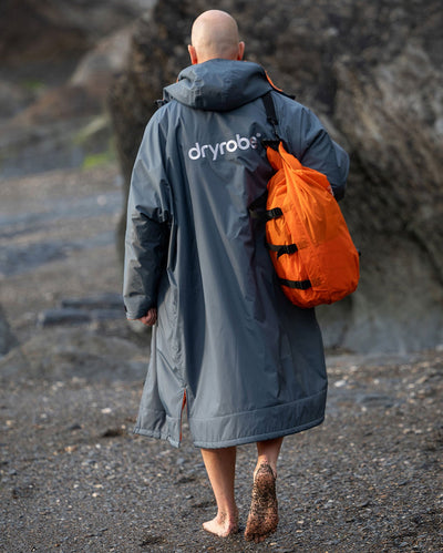 **MALE** Man walking along beach with back to the camera, wearing dryrobe Advance Long Sleeve change robe in Dark Grey Orange, carrying compression bag 