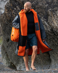 **MALE** Man stood in front of rocks on the beach, wearing dryrobe Advance Long Sleeve change poncho in Dark Grey Orange, carrying compression bag 