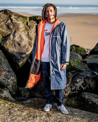 **MALE** Man stood on beach in front of sea and rocks, wearing dryrobe Advance Long Sleeve change poncho in Dark Grey Orange