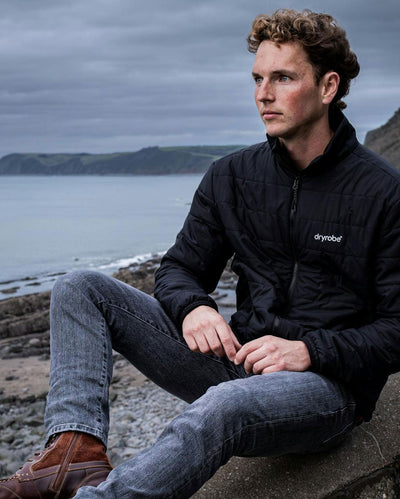 Man sat on rocks by the sea, wearing dryrobe Mid-Layer Jacket in Black 