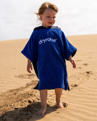 Toddler stood in sand on beach, wearing Kids Organic Towel dryrobe in Blue Grey