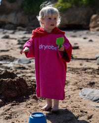 Child playing with toys on the beach wearing Kids Organic Towel dryrobe in Pink Red