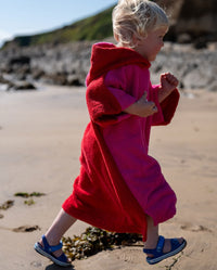 Child running along the beach, wearing Kids Organic Towel dryrobe in Pink Red