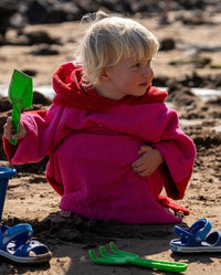 Toddler sitting on the beach holding a spade, wearing Kids Organic Towel dryrobe in Pink Red