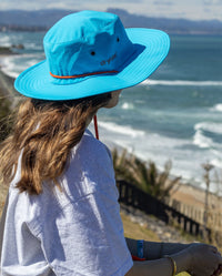 Woman sat side on in front of the sea, wearing dryrobe Quick Dry Brim Hat in Blue