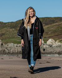 1|Woman walking towards camera on beach, wearing Black Grey dryrobe® Advance Short Sleeve