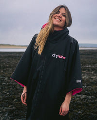 1|Woman stood smiling on a beach, wearing Black Pink dryrobe® Advance Short Sleeve zipped up 