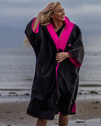 1|Woman stood in front of the sea, wearing Black Pink dryrobe® Advance Short Sleeve unzipped
