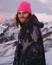 Man stood in mountains wearing Black Camo Pink dryrobe® Advance and Pink dryrobe® Eco beanie