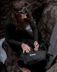 Woman in diving kit sat on rocks using the front pocket of the dryrobe® Eco Compression Backpack