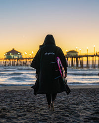 Woman carrying surfboard on Huntington Beach, wearing dryrobe® Lite