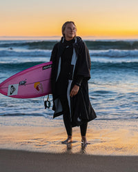 Woman stood in front of the ocean, carrying surfboard wearing dryrobe® Lite