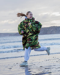 1|Girl smiling running along the shoreline, wearing Camo Grey dryrobe® Advance Kids Long Sleeve