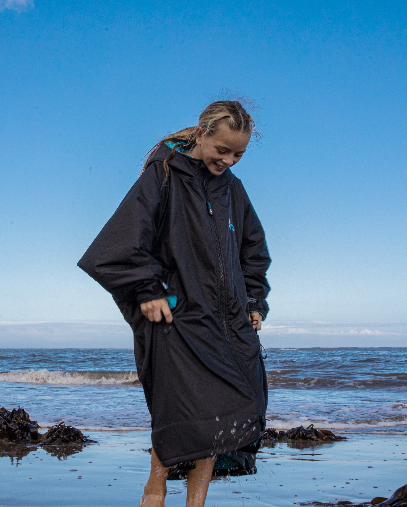 1|Girl stood on beach in front of the sea, wearing Black Blue dryrobe® Advance Kids Long Sleeve