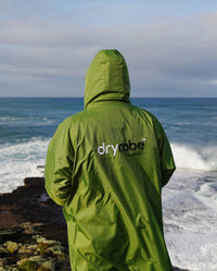 *MALE* Person stood facing the sea, wearing Forest Green dryrobe® Advance Long Sleeve with hood up