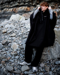 *MALE* sat on a rock on the beach, pulling hood up on Black dryrobe® Waterproof Poncho 