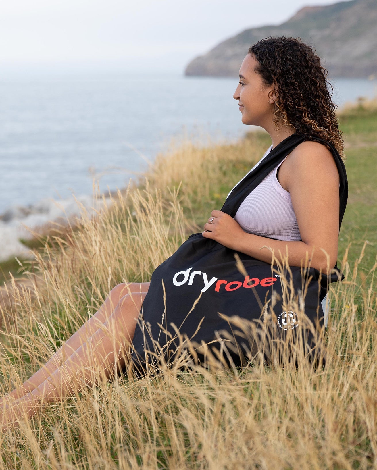 Woman sat on grassy cliffs above the sea holding dryrobe® Tote Bag